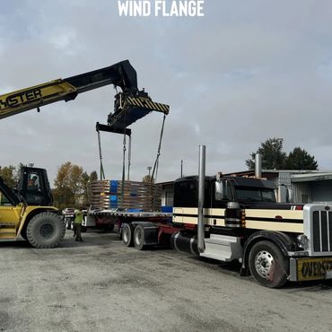 A crane lifts a large wind flange onto a flatbed truck labeled oversize load.