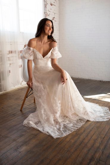 A woman in an elegant off-shoulder lace wedding dress sitting on a chair.
