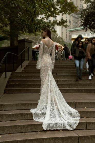 A woman in an elegant lace gown stands on outdoor steps in an urban setting.