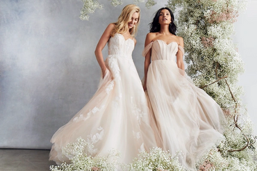 Two women in elegant, flowing wedding dresses surrounded by delicate white flowers.