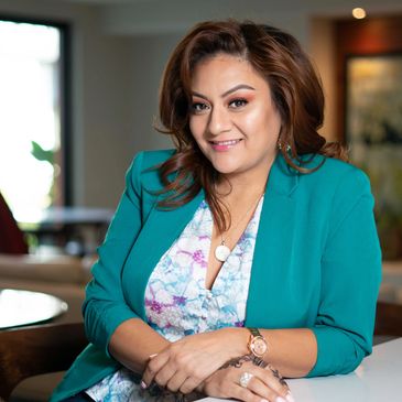 A Latina woman in a teal blazer and pastel blouse smiles at the camera