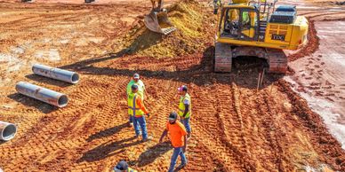 Construction workers discuss plans on a muddy site with heavy machinery.