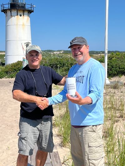 Two men shaking hands near a lighthouse on a sunny day.