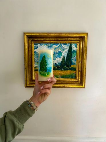 Hand holding a candle painted with a landscape in front of a framed painting of the same scene.