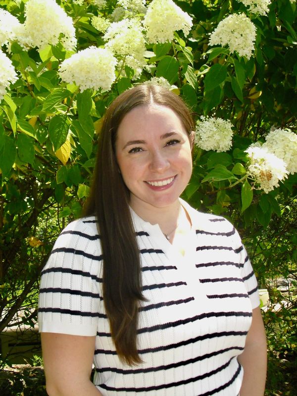 Smiling woman in a striped shirt stands in front of blooming white flowers.