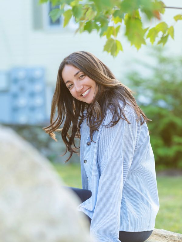 Smiling woman in a light blue shirt outdoors with green foliage.