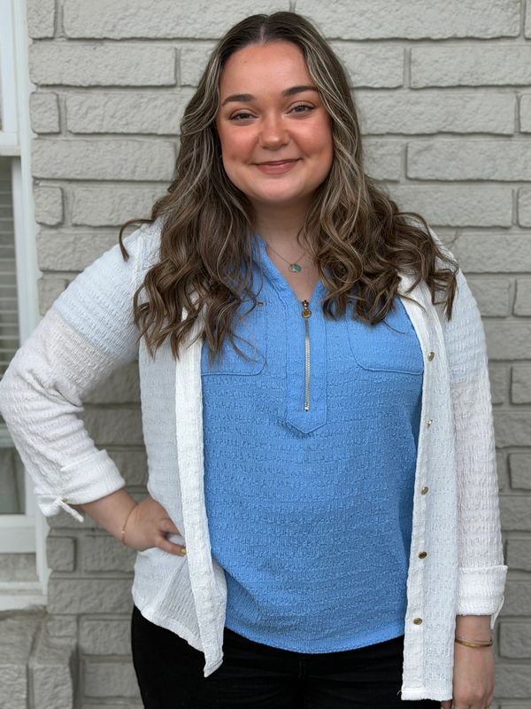 Young woman with wavy hair smiles in front of a gray brick wall.