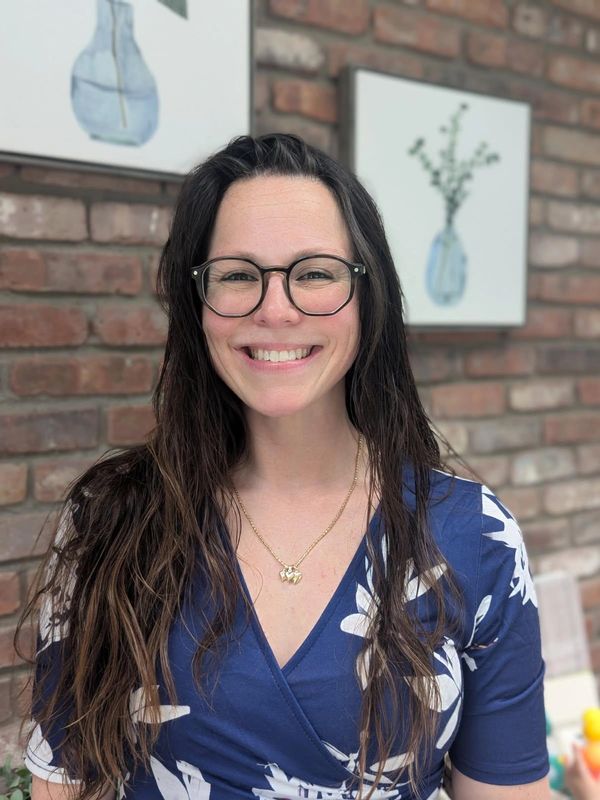Smiling woman with glasses in a blue floral dress against a brick wall.