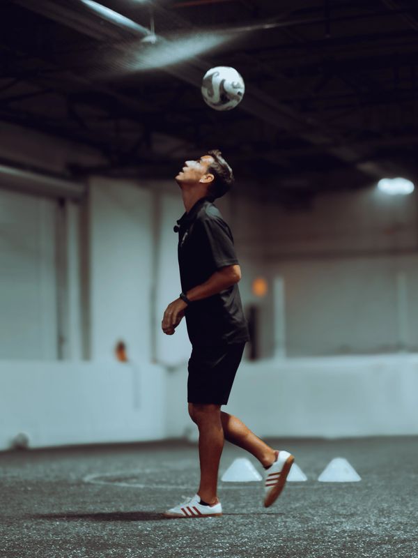 Man in black outfit balancing a soccer ball on his head indoors.