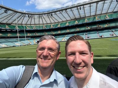 Two men smiling for a selfie inside a large, empty sports stadium.