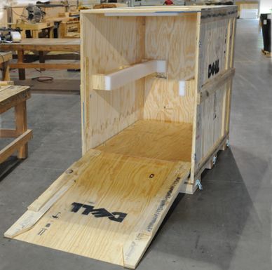 An open wooden crate with a ramp, featuring a Dell logo, situated in a workshop with various tools