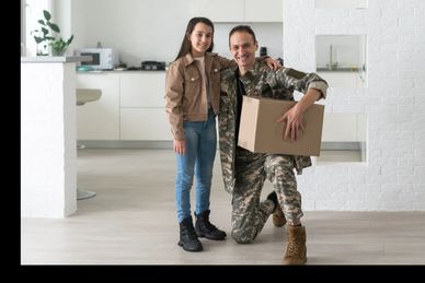 A soldier in uniform kneels with a young girl in a modern kitchen holding a cardboard box.