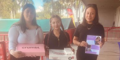 Three women holding donut boxes and a flyer outdoors with palm trees in the background.