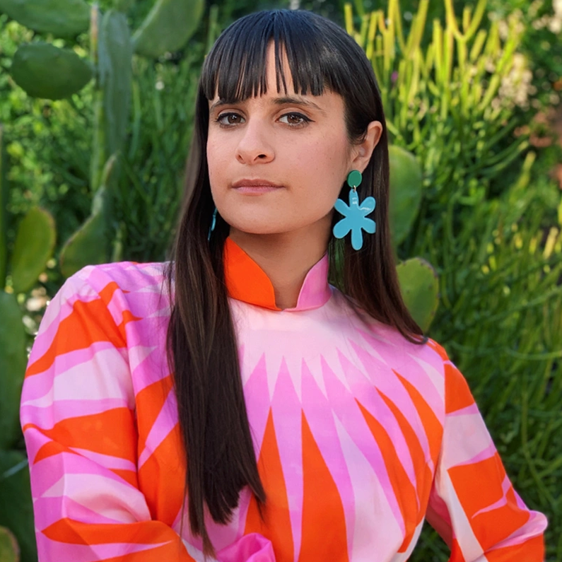 Woman with long hair wearing colorful pink and orange dress with blue flower earrings.