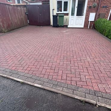 A clean, patterned brick driveway in front of a house with a garden and bins.