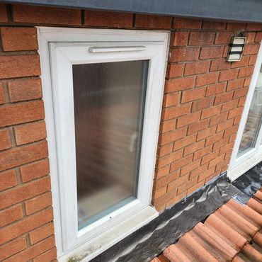 A small fogged-up white window on a brick wall above a tiled roof.