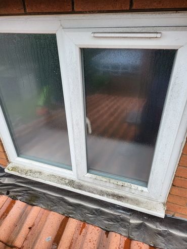 A foggy window with condensation and dirt on the frame above a tiled roof.