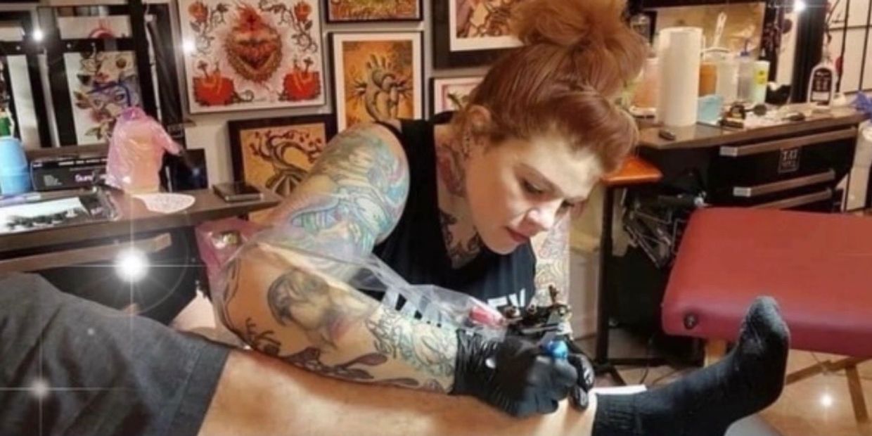 Tattoo artist with colorful arm tattoos working on a client's leg in a tattoo studio.