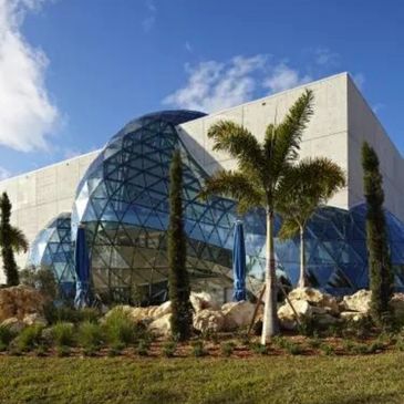 Modern building with glass dome and palm trees under blue sky.