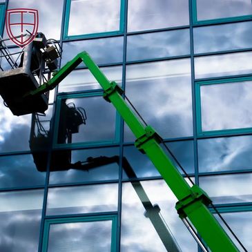 Green boom lift working on reflective glass building with cloudy sky.