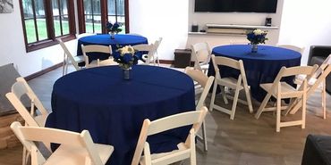 Indoor party setup with blue tablecloths and white chairs around round tables.