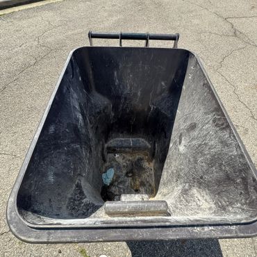 Empty black wheelbarrow on a sunny street with a single foot visible.