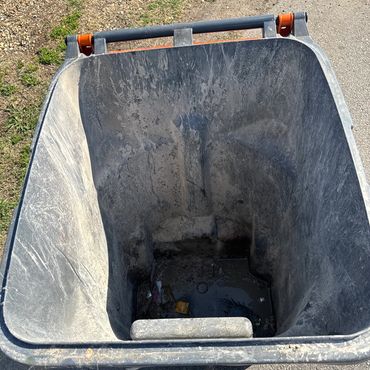 Empty, dirty black trash bin on a sunny outdoor surface.