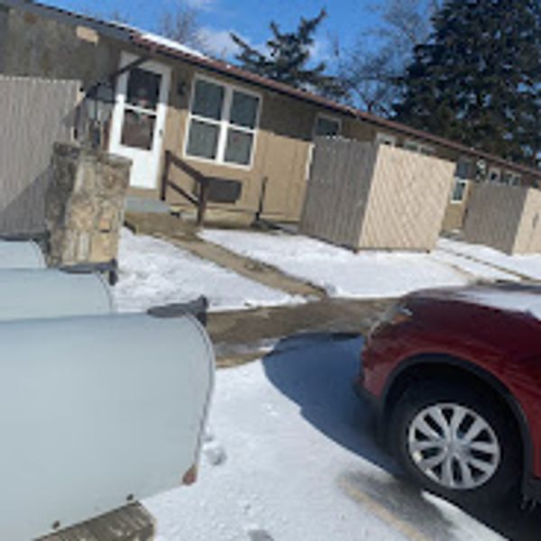 Snow-covered driveway with a red car and a small house in the background.