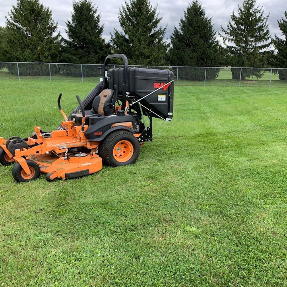 Orange zero-turn lawn mower on green grass with pine trees in background.