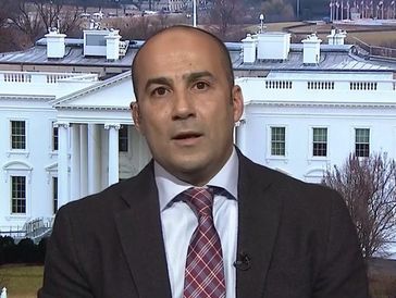 Man in a suit speaking with the White House in the background.