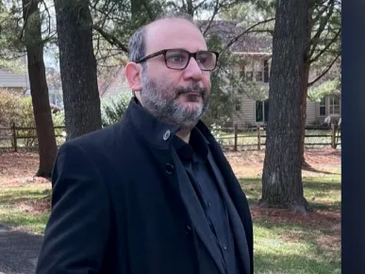 Man in black coat standing outdoors near trees and a house.