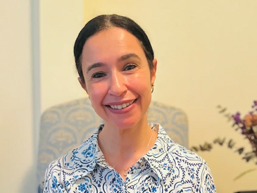 Smiling woman in a blue and white floral patterned shirt sitting indoors.