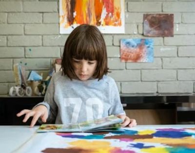 A young girl reading a book while sitting at a table with colorful paintings.