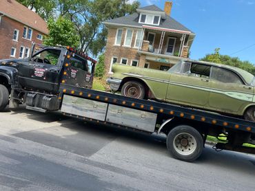 A black tow truck carrying an old green car on a sunny day.