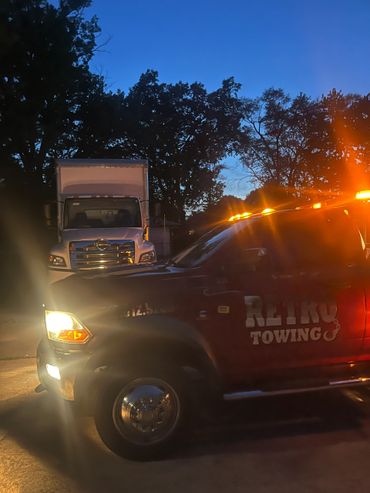 Tow truck with flashing lights at dusk near a parked delivery truck.