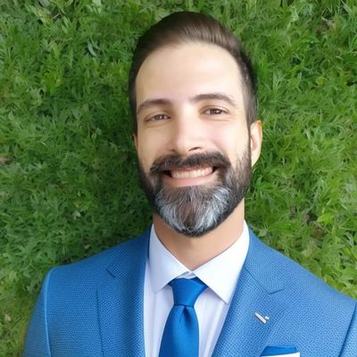 Smiling man with a beard in a blue suit against a green leafy background.