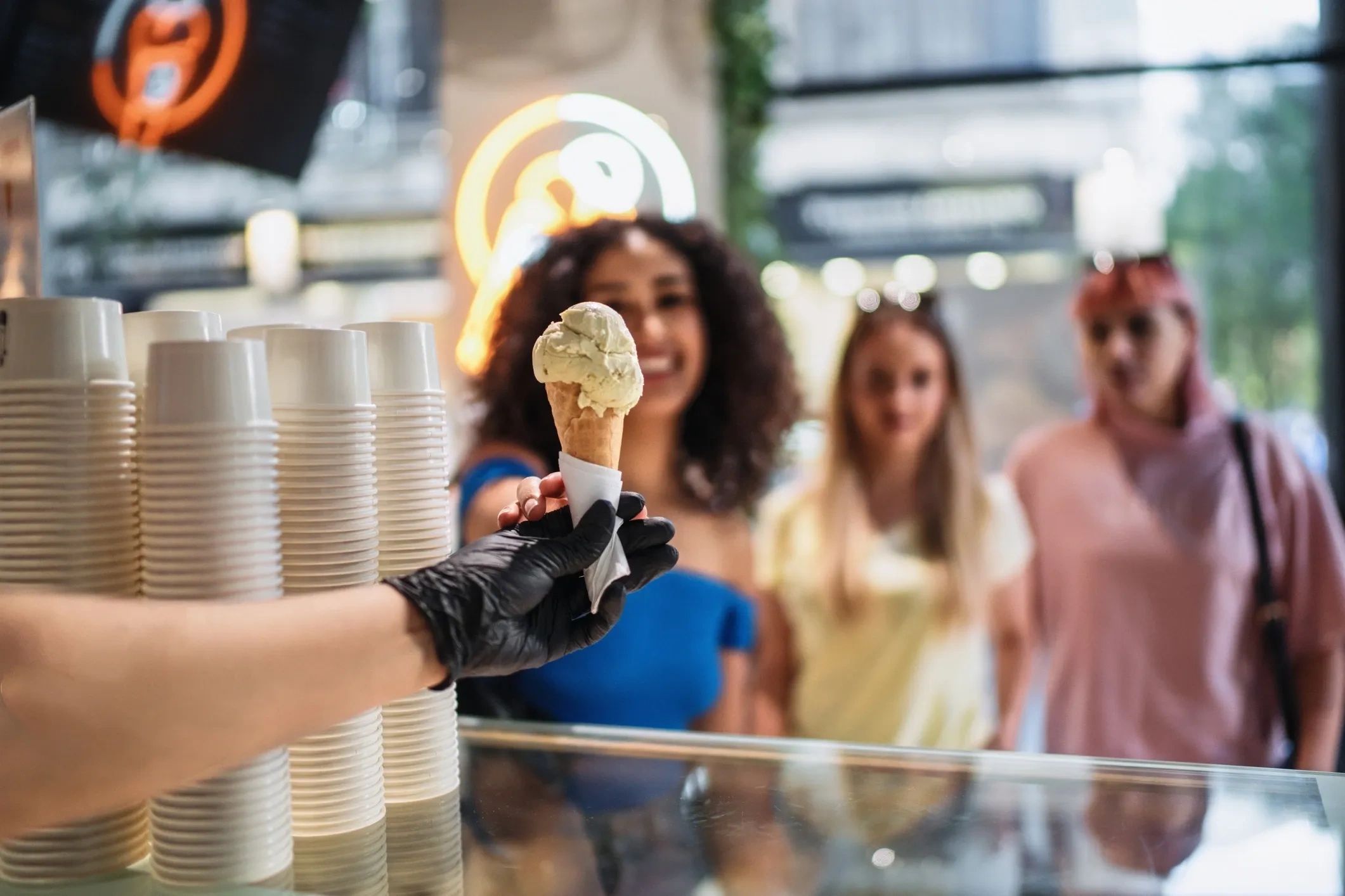 A hand wearing a black glove serves ice cream to three smiling women at a counter.