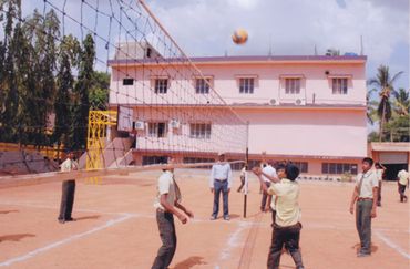 Students playing volleyball on a dusty outdoor court near a pink building.