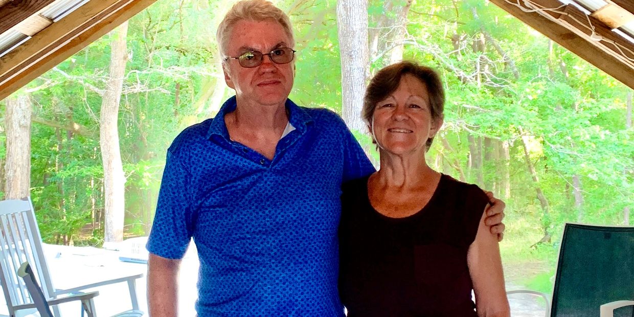A mature couple smiling under a wooden roof with forest background.