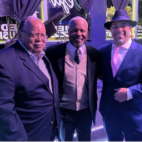 Three men dressed in suits posing happily indoors at a music venue.
