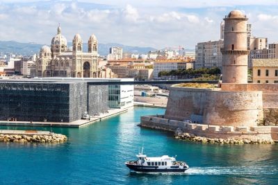 Boat sailing near historic fort and modern buildings in a coastal city.