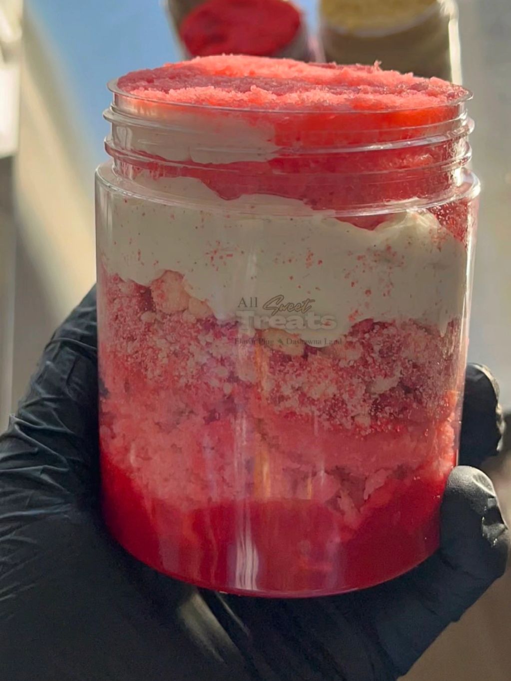A layered red and white dessert or treat in a clear jar held by a gloved hand.