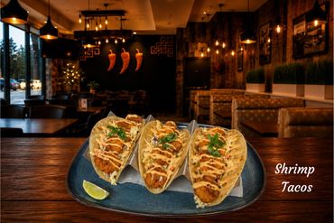 Three shrimp tacos served on a blue plate with lime, in a cozy restaurant setting.