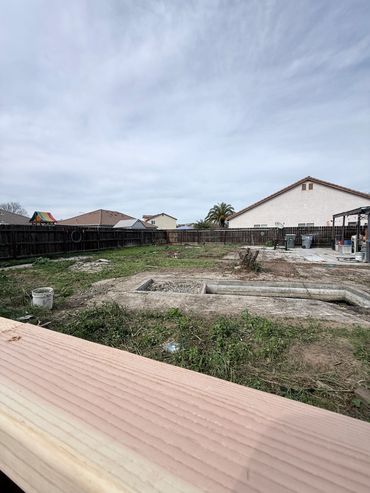 Empty backyard with old pool structure and wooden fence under a cloudy sky.
