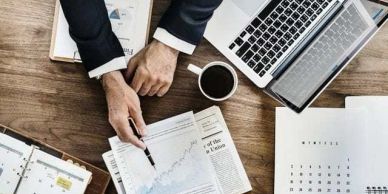 Person analyzing financial charts with a laptop and coffee on a wooden desk.