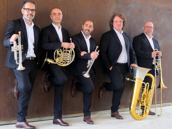 Five men in suits posing with brass instruments against a brown wall.