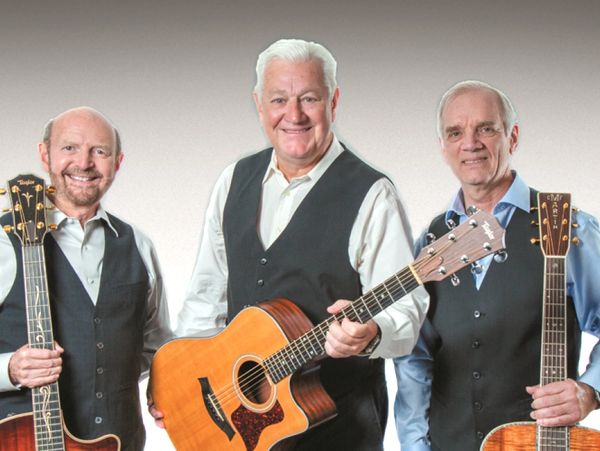 Three elderly men smiling with acoustic guitars, dressed in formal vests.