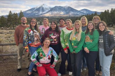 A group of people wearing festive holiday sweaters posing outdoors with mountains in the background.