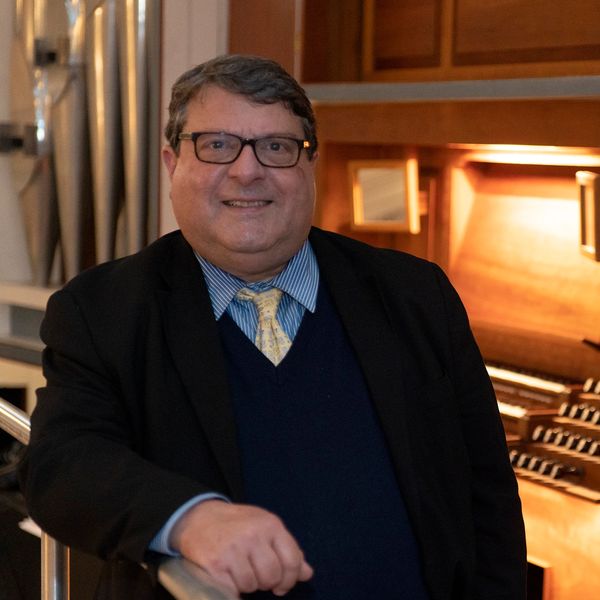 Man in glasses and suit standing by an organ.