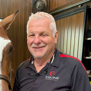 Smiling man with white hair posing next to a horse indoors.